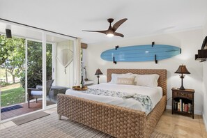 Ein Schlafzimmer mit Bett, Deckenventilator, Surfbrett an der Wand, Bücherregal und einer Schiebeglastür zum Garten.