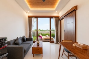 A modern living room with a sofa, coffee table, and dining table. There is a sliding glass door leading to a balcony with a view of greenery. - Aswindra Private Villa (Ubud)