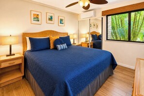 A bedroom with a large bed, wooden headboard, two bedside tables with lamps, a ceiling fan, and a window with a view of greenery. - Napili Point | Oceanfront 2 BR, Sleeps 8 | Car Incl. w/ 6+ Nights | NAP ML-4154 KBM (Lahaina)