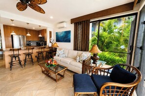 A modern living area with a sofa, coffee table, and wicker chairs, a kitchen with bar stools, and a large window with a view of greenery. - Napili Point | Oceanfront 3 BR, Sleeps 12 | Car Incl. w/ 6+ Nights | NAP ML-4157 by KBM (Lahaina)