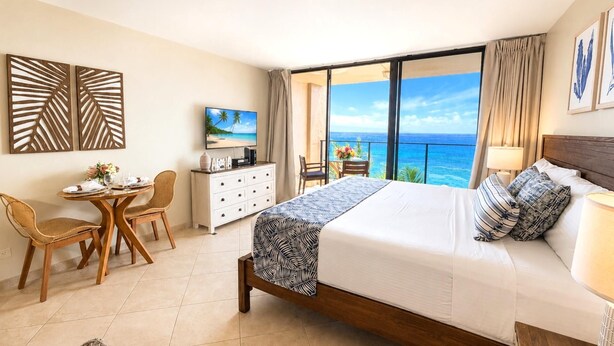 A hotel room with a bed, a desk, a chair, a TV, and a balcony with a view of the ocean. - Mahana Villas | Oceanfront 3 BR Sleeps 10 | Car Incl. w/ 6+ Nights | MAH ML-4160 by KBM (Lahaina)