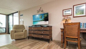 A living room with a sofa, a TV, a wicker side table, a chair, and a bookshelf. - Mahana Villas | Oceanfront 3 BR Sleeps 10 | Car Incl. w/ 6+ Nights | MAH ML-4160 by KBM (Lahaina)