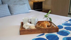 En pænt redt seng med en bakke croissanter, to hvide kopper og et sammenrullet håndklæde.