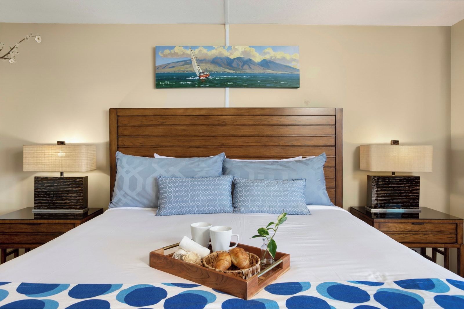A hotel room with a wooden headboard, two nightstands with lamps, a bed with a blue and white patterned bedspread, a tray with breakfast items, and a painting of a sailboat on the wall.