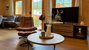 En inredning med träpanel, en soffa, ett runt bord med ett ljus, en tv och ett fönster med utsikt över grönområden.
