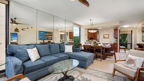 A modern living room with a sectional sofa, a glass coffee table, and a dining area with wooden furniture. - Mahana Villas | Oceanfront 5 BR Sleeps 13 | Car Incl. w/ 6+ Nights | MAH ML-4169 by KBM (Lahaina)