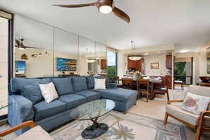 A modern living room with a sectional sofa, a glass coffee table, and a dining area with wooden furniture. - Mahana Villas | Oceanfront 5 BR Sleeps 13 | Car Incl. w/ 6+ Nights | MAH ML-4169 by KBM (Lahaina)