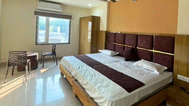 A hotel room with a large bed, a desk, a chair, a window, and a ceiling fan. - Kashika Inn and Comfort Stays (Varanasi)