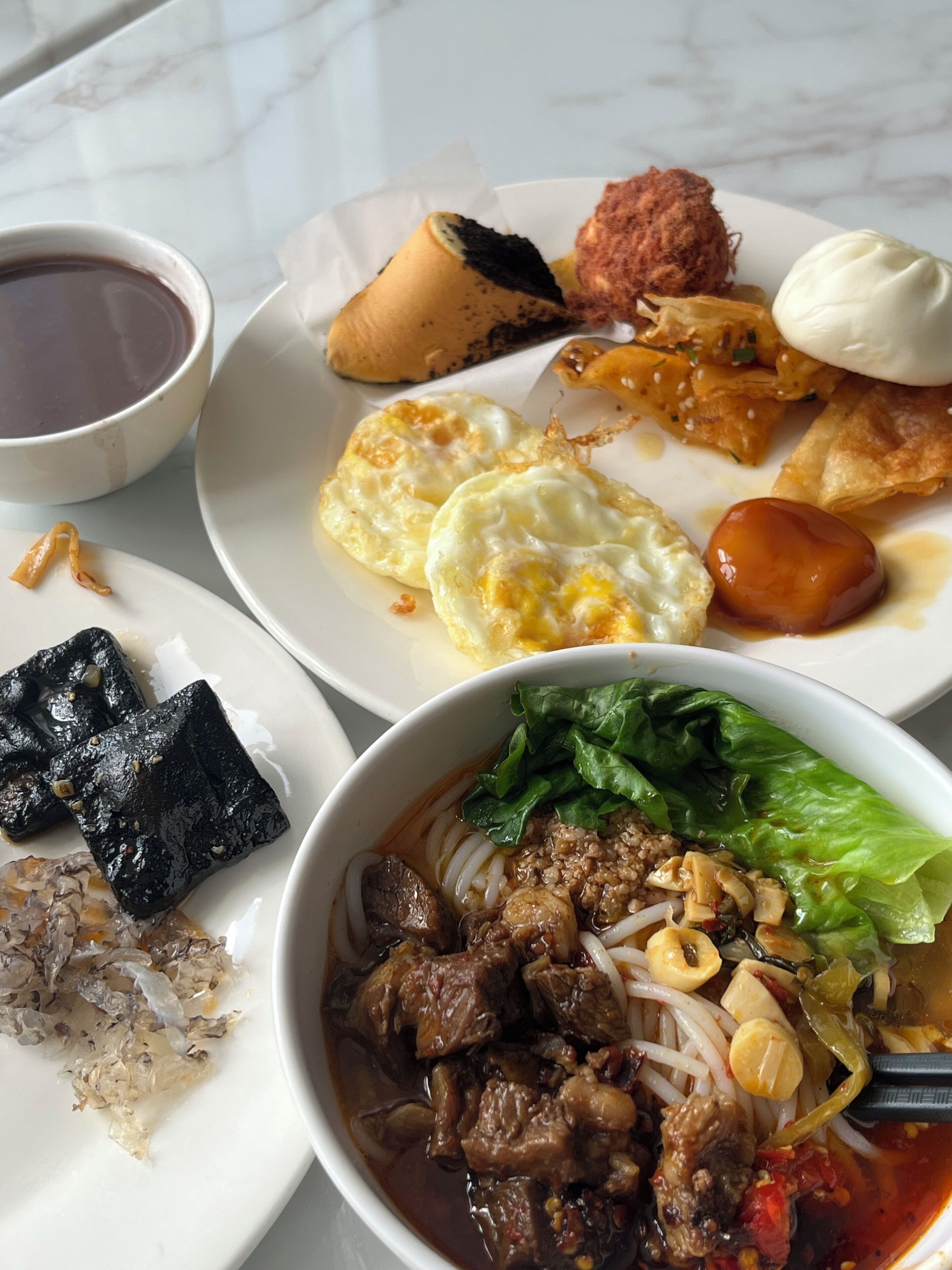 A variety of dishes including a bowl of soup with meat and vegetables, a plate with fried items and a bun, and a cup of a dark beverage.