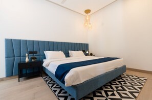 Un dormitorio con cabecera azul, cama con ropa de cama blanca y azul, alfombra estampada en blanco y negro, y una mesita de noche con una lámpara.