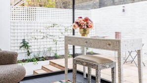 Une chambre moderne avec un tapis à motifs, une table en bois ornée d’un vase de fleurs et une vue sur un bâtiment en briques, visible à travers une fenêtre.