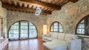 Quarto com paredes de pedra, teto de madeira, uma grande janela em arco, um sofá e piso ladrilhado.