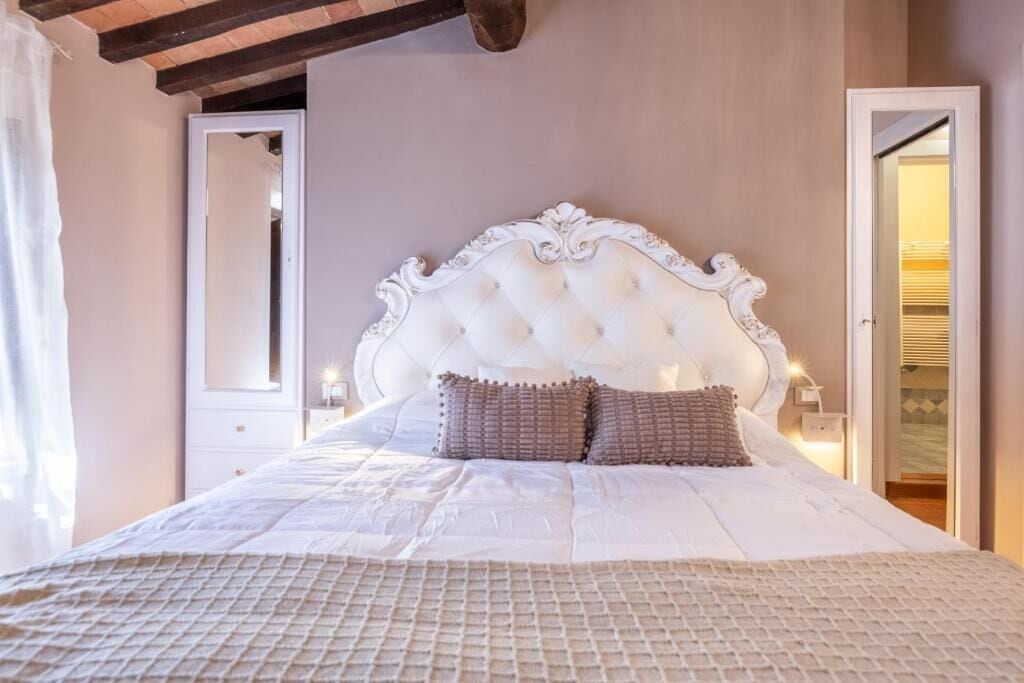 Un dormitorio con cabecera capitonada, una cama grande con colcha acolchada, un espejo y una mesita de noche con lámpara.