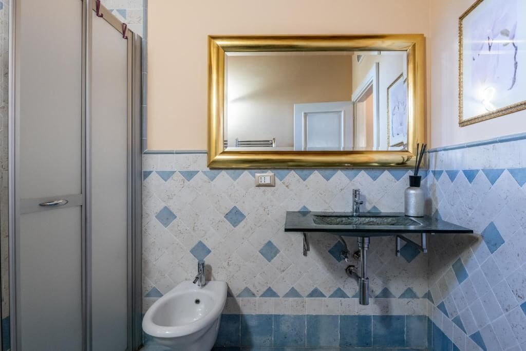 A bathroom with a bidet, a sink with a glass countertop, a gold-framed mirror, and a tiled wall with a diamond pattern.