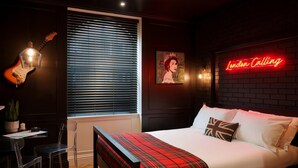 A hotel room with a bed, a neon sign, a guitar, a window with blinds, and a painting on the wall. - Guestready - Camden Studios (London)