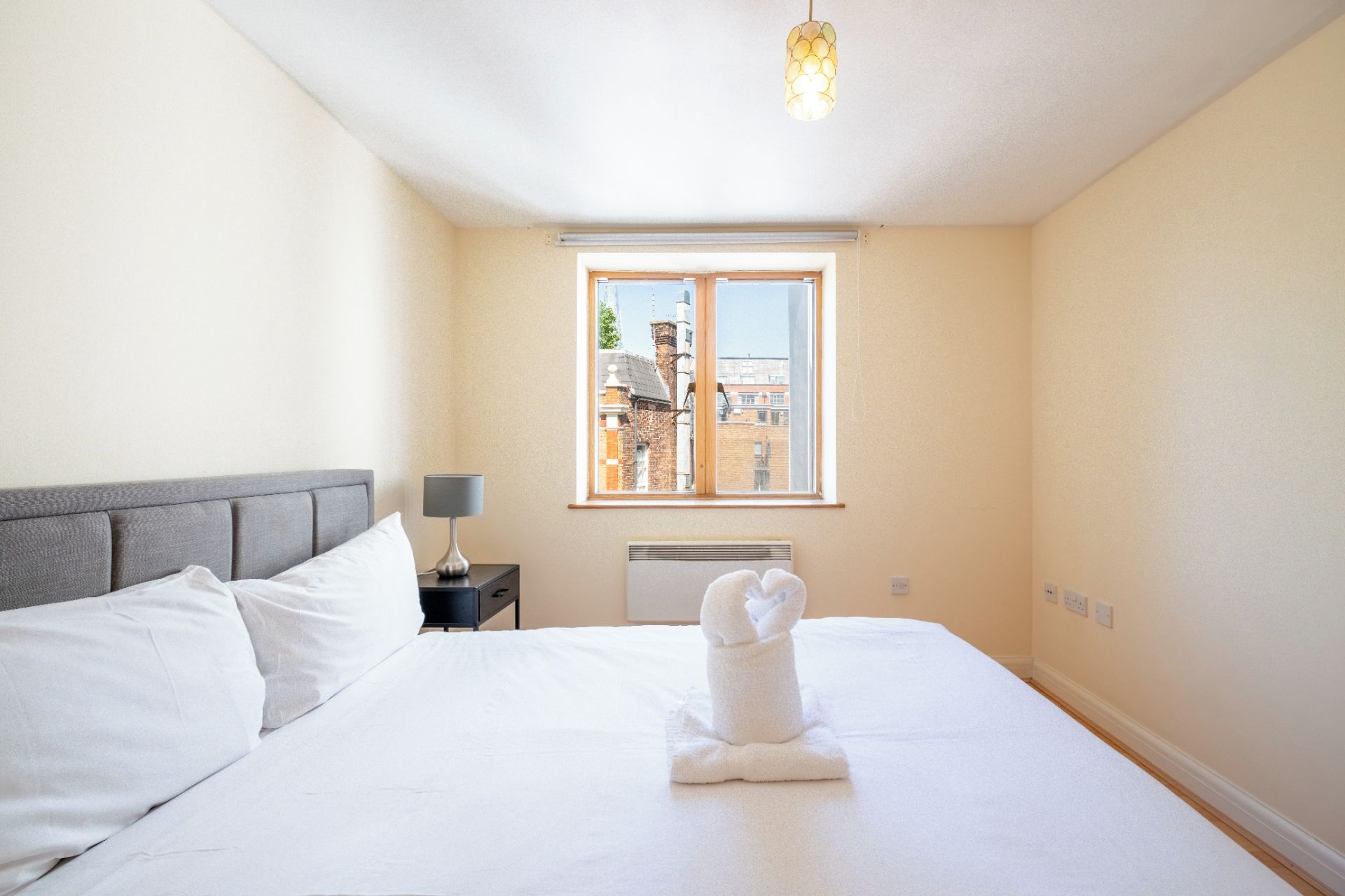 Ett hotellrum med en säng, ett nattduksbord, en lampa, ett fönster med utsikt över byggnader och en konstnärlig dekoration av handdukar på sängen.