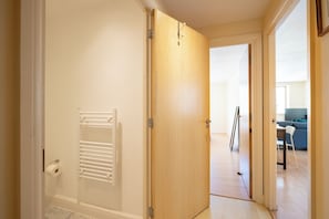 Un couloir mène à une pièce dotée d’une porte en bois, d’un radiateur, et on aperçoit un coin salon avec un canapé et une table.