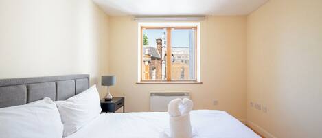 Une chambre d’hôtel avec un lit, une table de chevet, une lampe, une fenêtre donnant sur des immeubles et une décoration artistique réalisée avec des serviettes sur le lit.