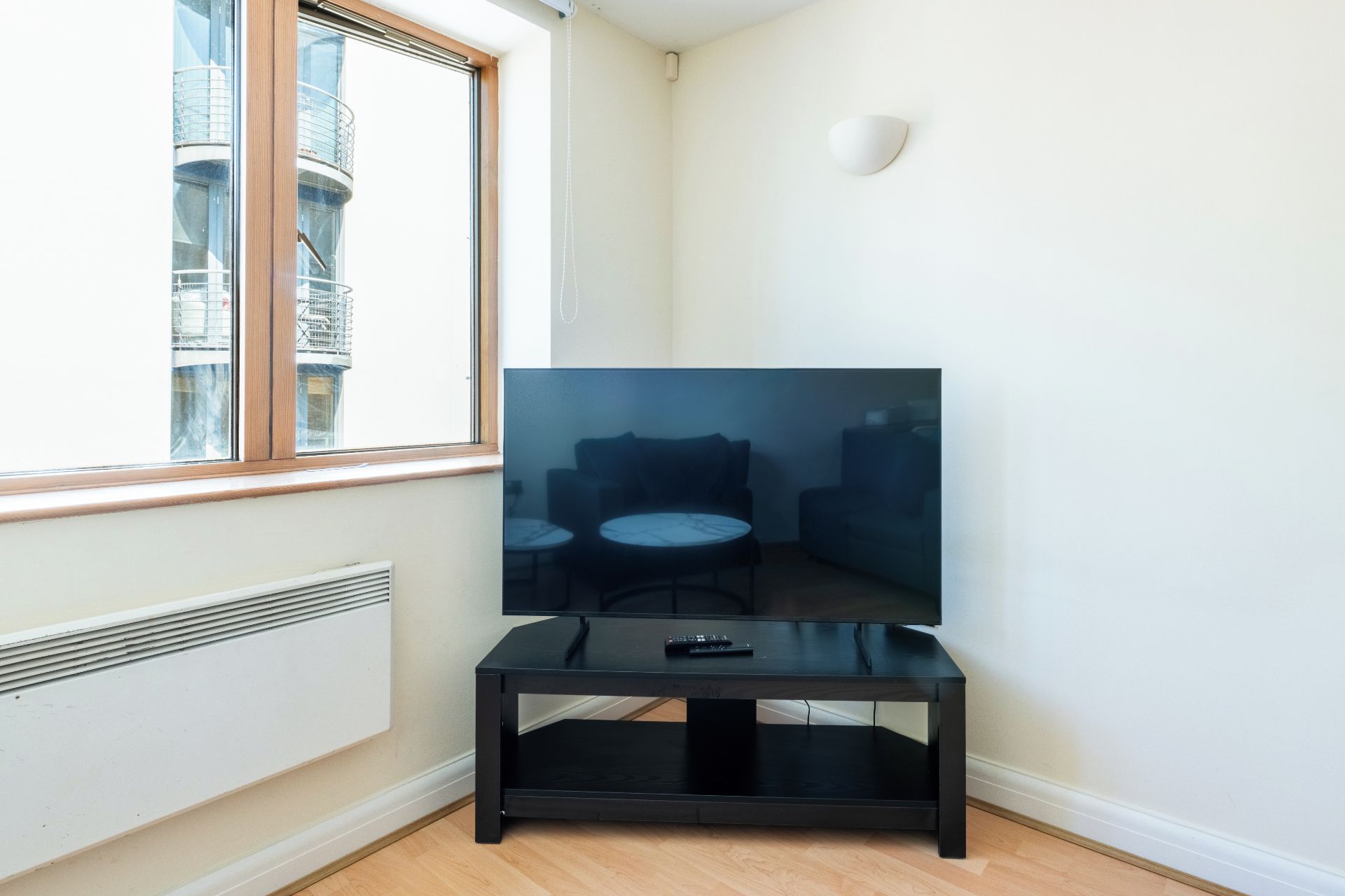 A corner room with a window, a radiator, a black TV stand, and a remote control on it.