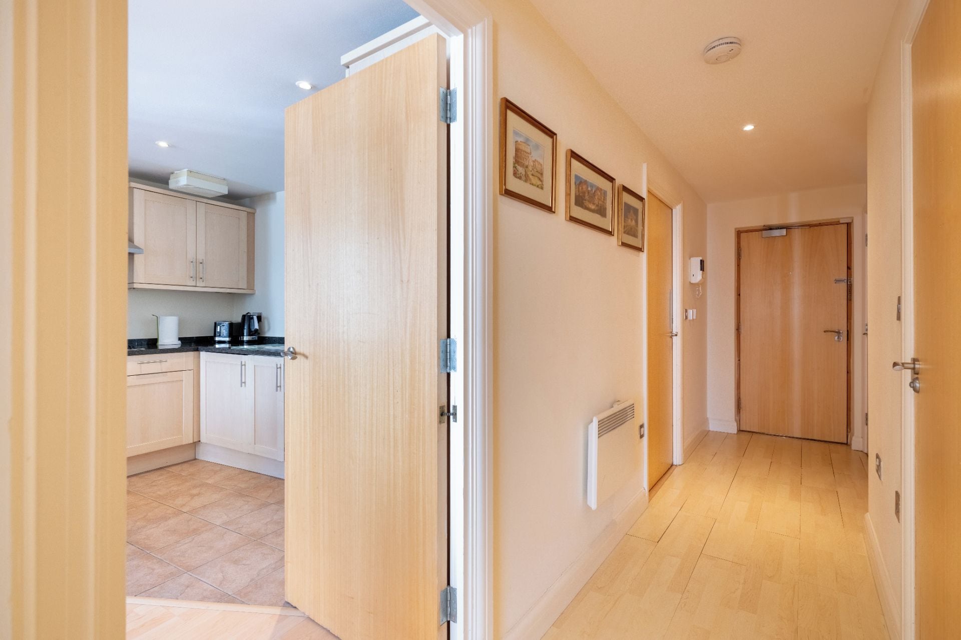 A hallway with a kitchen area, wooden flooring, and a door leading to another room.