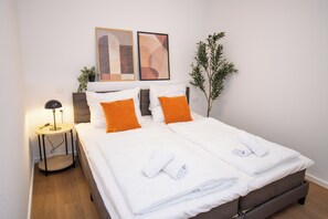 Ein ordentlich bezogenes Doppelbett mit weißer und orangefarbener Bettwäsche, ein Nachttisch mit Lampe sowie zwei abstrakte Gemälde an der Wand.