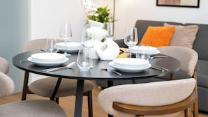 Une table à manger dressée avec des assiettes, des verres et des couverts, entourée de chaises, dans une pièce où se trouvent un canapé et une plante.