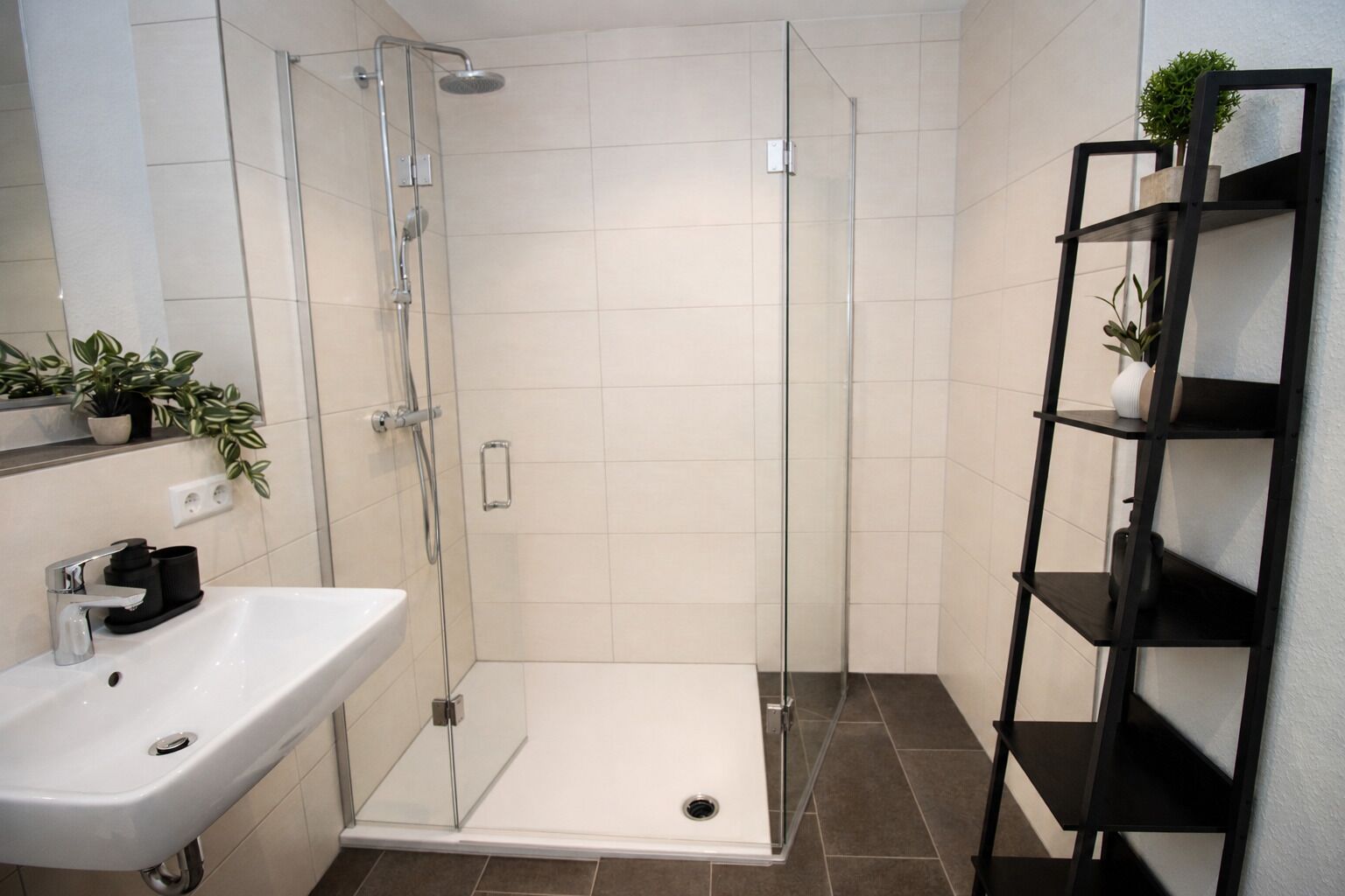 Ein modernes Badezimmer mit einer ein Glasfach raumteilenden Dusche, einem weißen Waschbecken und einer schwarzen Regaleinrichtung mit Pflanzen.