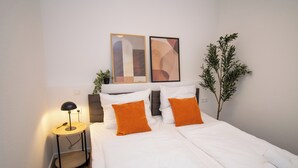 Un lit bien fait, avec une literie blanche et des oreillers orange, une lampe de chevet et une œuvre d’art abstraite accrochée au mur.