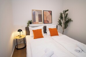 Ein ordentlich bezogenes Bett mit weißer Bettwäsche und orangefarbenen Kissen, eine Nachttischlampe und abstrakte Kunst an der Wand.