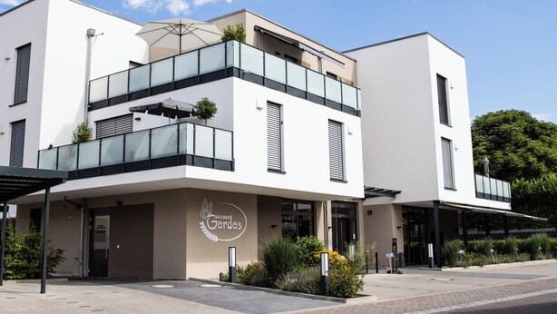 Un bâtiment moderne doté d’un balcon en verre, d’une enseigne indiquant « Bäckerei Gerdes » et d’une entrée abritée.