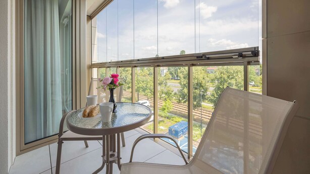 A balcony with a table, chairs, a vase with flowers, and a view of greenery and buildings. - Bright Apartment with a Balcony | Parking (Warszawa)