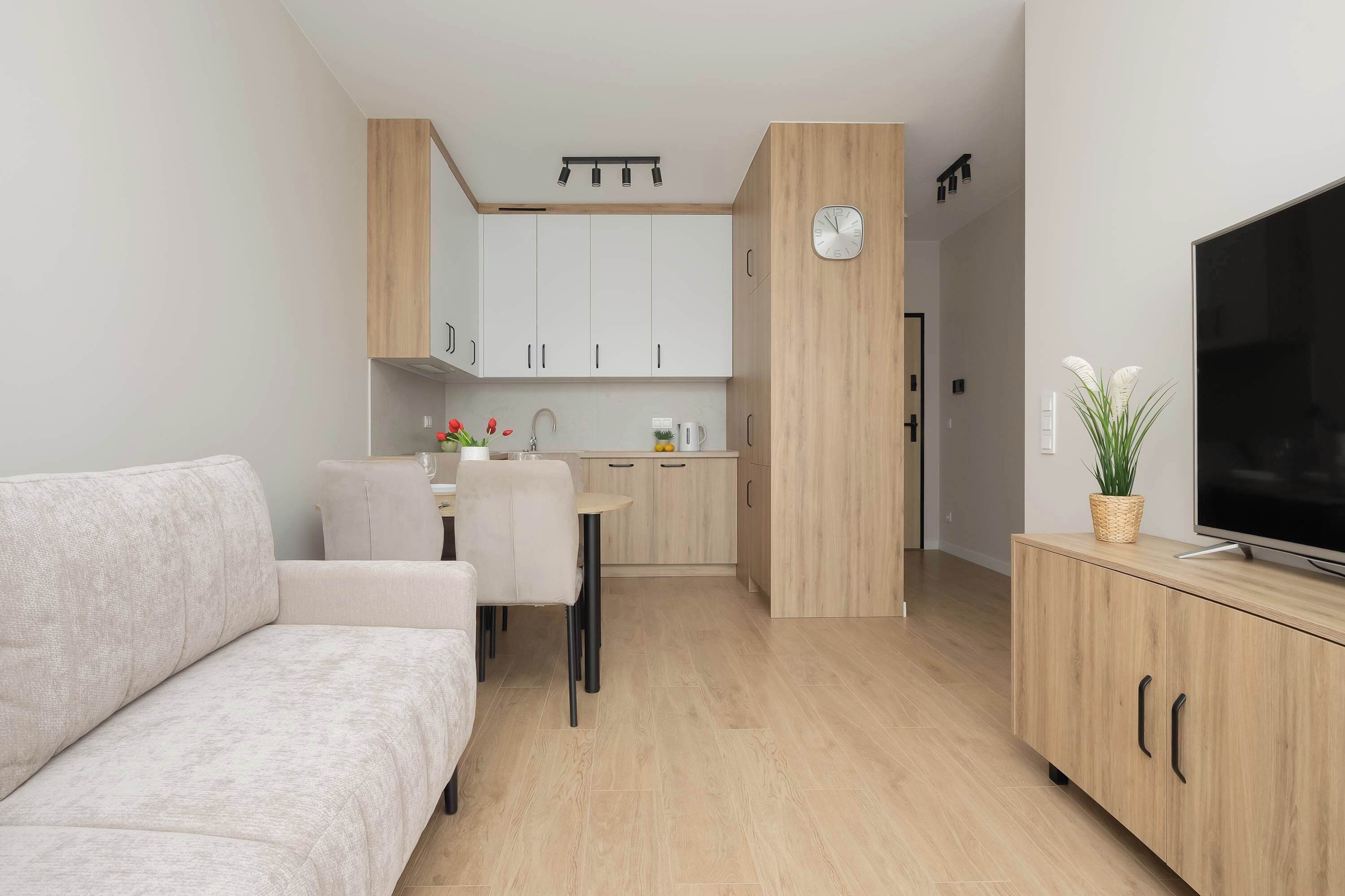Una sala de estar moderna con un sofá, una zona de comedor con sillas, una cocina con armarios, un reloj y un televisor sobre un mueble de madera.