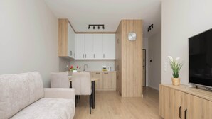 A modern living room with a sofa, a dining area with chairs, a kitchen with cabinets, a clock, and a TV on a wooden cabinet. - Bright Apartment with a Balcony | Parking (Warszawa)