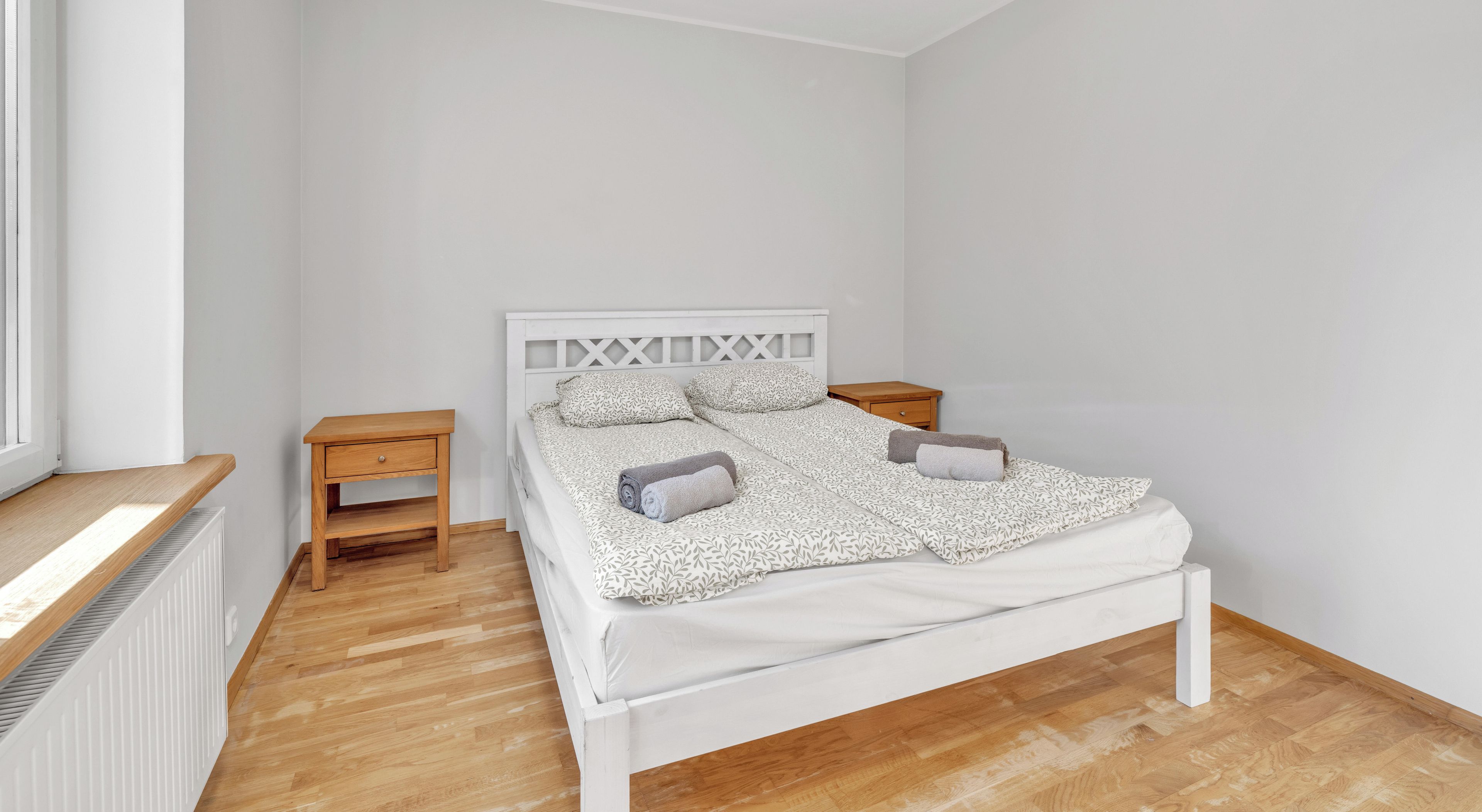 Een slaapkamer met een wit bed, twee nachtkastjes, een raam en een radiator.