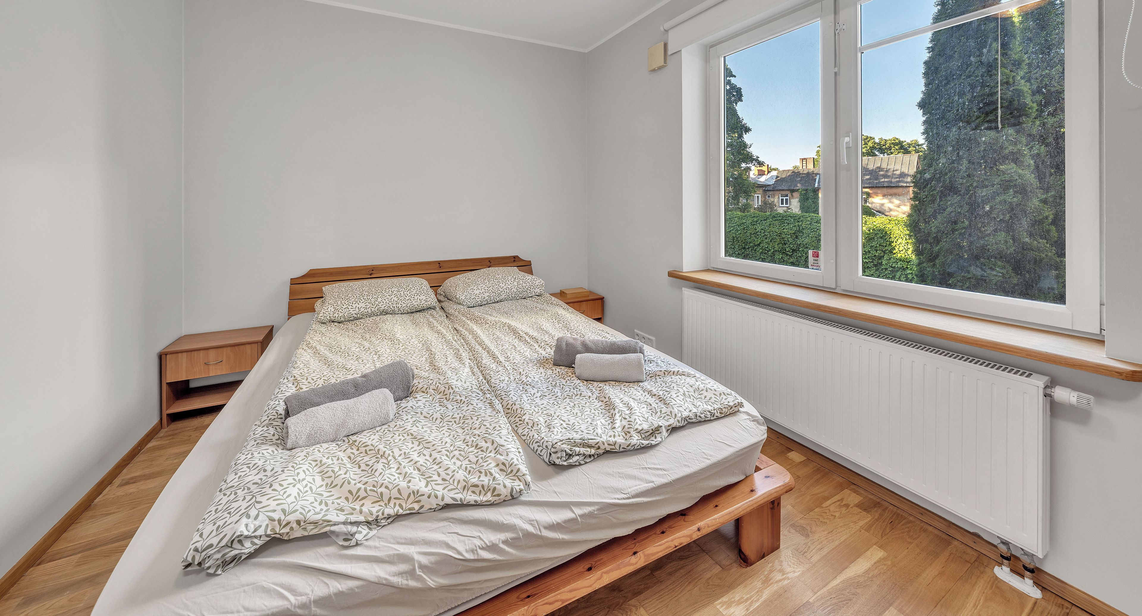 Een enkel bed met een houten hoofdbord, een houten bank voeteneind en een nachtkastje, plus een raam met uitzicht op groen en gebouwen.
