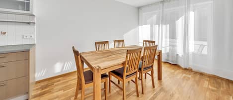 Un coin repas avec une table et des chaises en bois, une cuisine équipée de placards et une fenêtre ornée de rideaux légers.