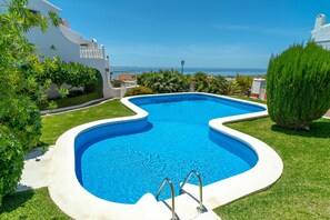 Ein Pool mit ungewöhnlicher Form, umgeben von gepflegtem Rasen und Grünflächen, mit Blick auf das Meer im Hintergrund.