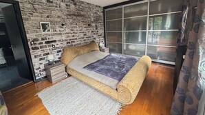 A bedroom with a brick wall, a bed with patterned bedding, a bedside table with a lamp, and a large sliding glass door. - Beautiful house (Bernay)