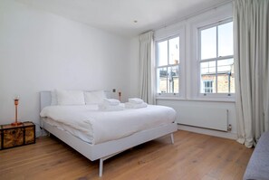 Une chambre à coucher avec un lit blanc, un plancher en bois et une fenêtre avec des rideaux.