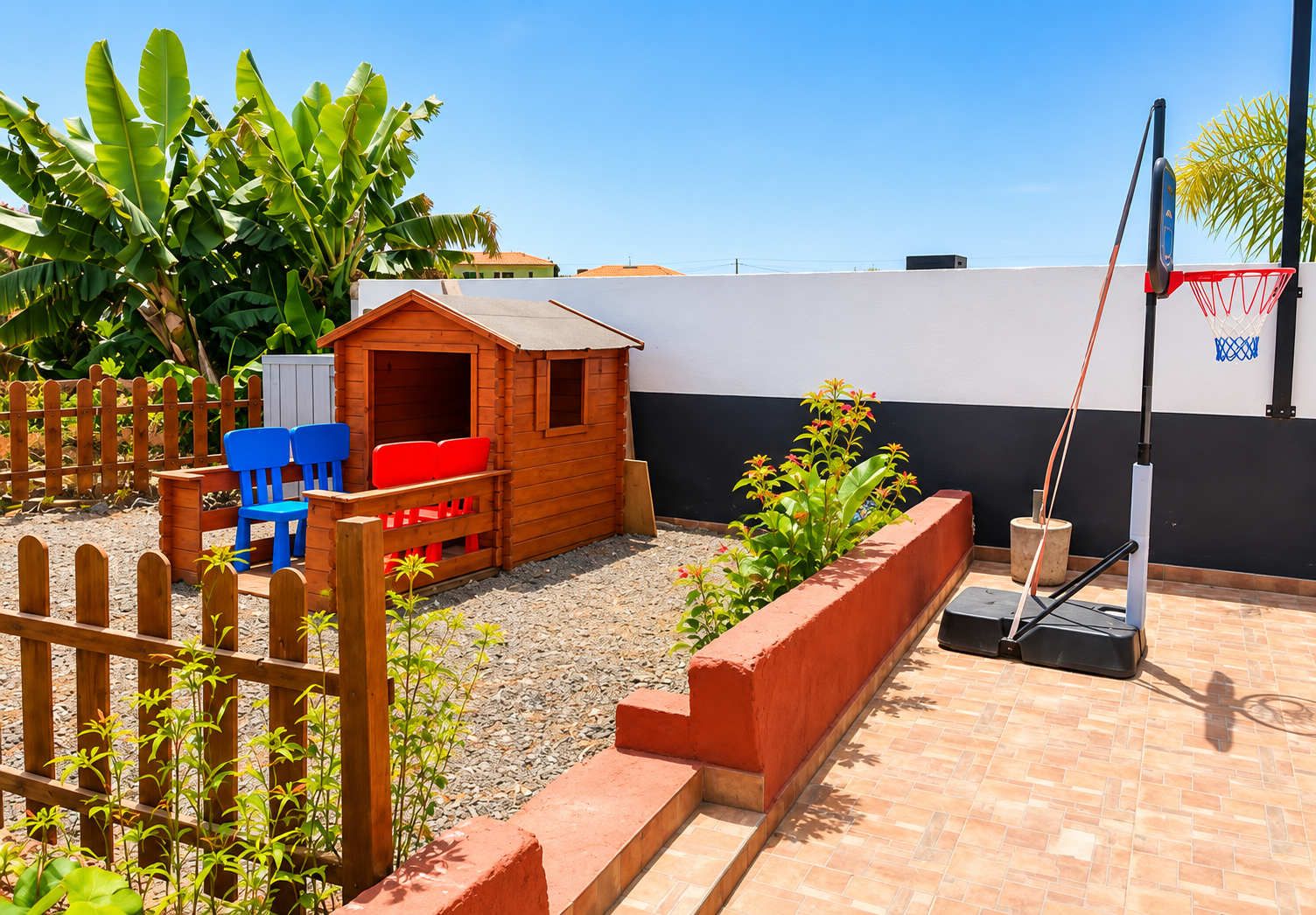 A children's play area with a wooden playhouse, blue and red chairs, a basketball hoop, and a garden with plants and a wooden fence.