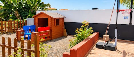 A children's play area with a wooden playhouse, blue and red chairs, a basketball hoop, and a garden with plants and a wooden fence.