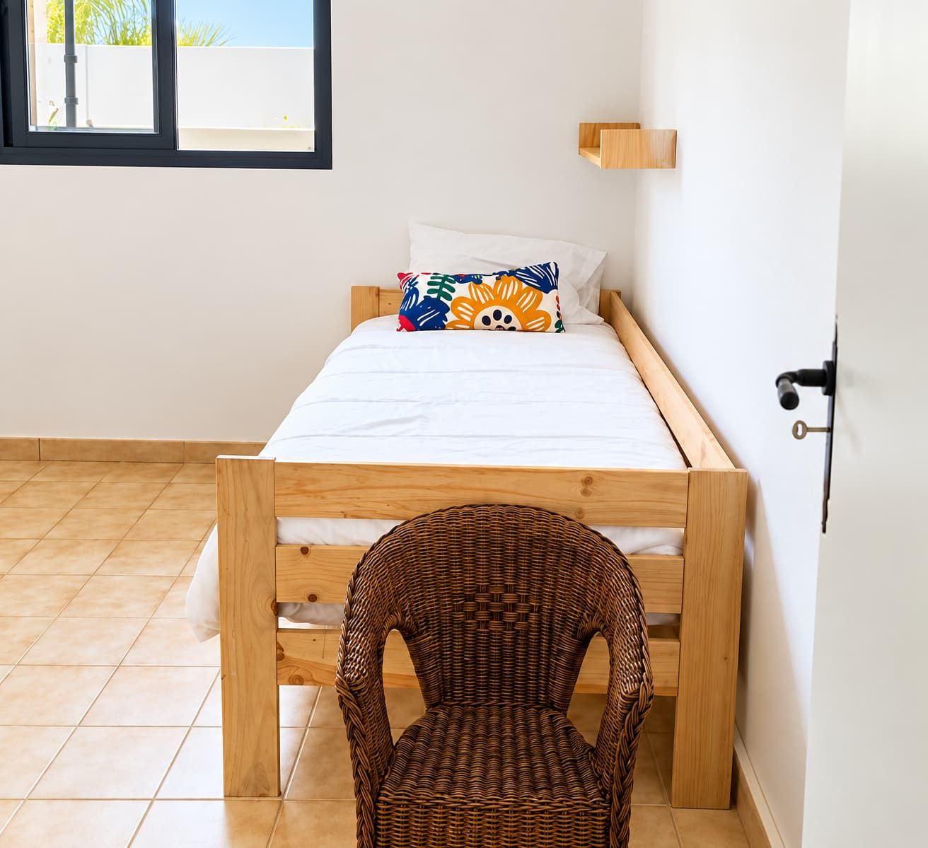 Une petite chambre avec un lit simple, un cadre en bois, une chaise en osier et une fenêtre donnant sur l’extérieur.