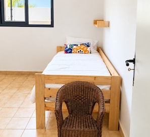 A small, single-bed room with a wooden frame, a wicker chair, and a window with a view of the outside. - Tacoronte House: Garden, BBQ, Parking (Tacoronte)