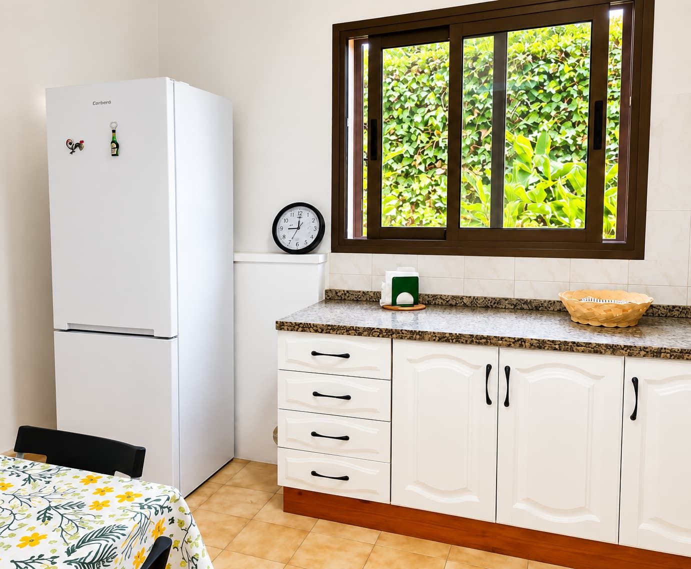 A kitchen with a white refrigerator, a clock, a window with a view of greenery, and a table with a floral tablecloth.