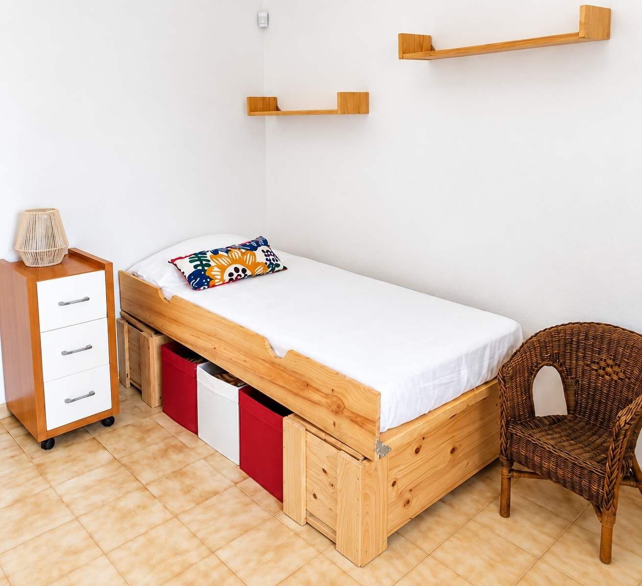 Una cama individual con estructura de madera, una mesita de noche pequeña, una silla y un mueble de almacenamiento rojo.