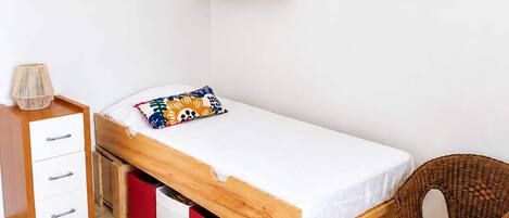 Una cama individual con estructura de madera, una mesita de noche pequeña, una silla y un mueble de almacenamiento rojo.