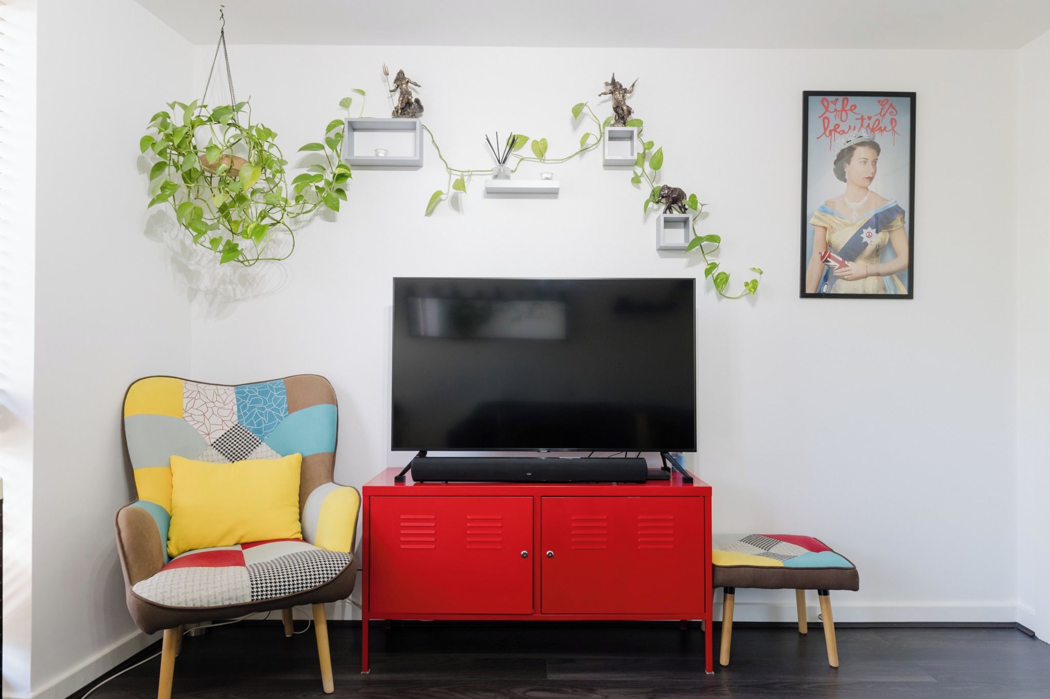 Una sala de estar con televisor, un mueble rojo, un sillón colorido, un banco y plantas decorativas.