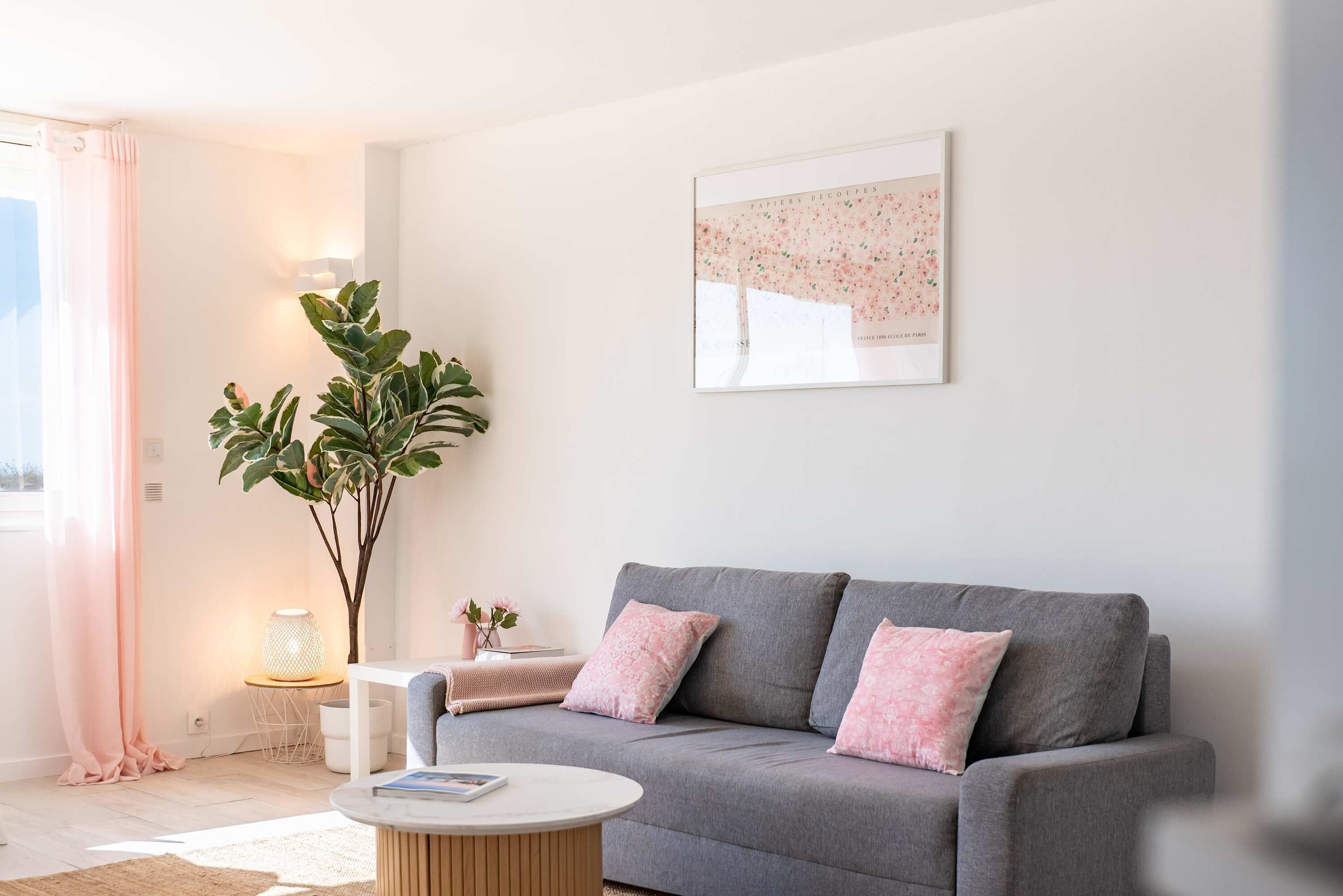 Een moderne zithoek met een grijze bank, een ronde salontafel, een potplant, een wandlamp en een ingelijst schilderij.