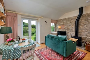 A cozy living room with a stone wall, a green sofa, a dining table with a polka dot tablecloth, and a view of greenery outside. - Mole End (Wass)