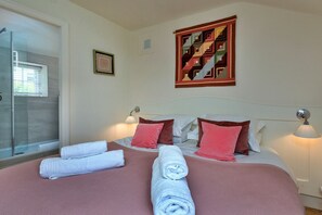Habitación de hotel con cama, dos lámparas de noche, una obra de arte enmarcada, un ventanal pequeño y un box de ducha de cristal.